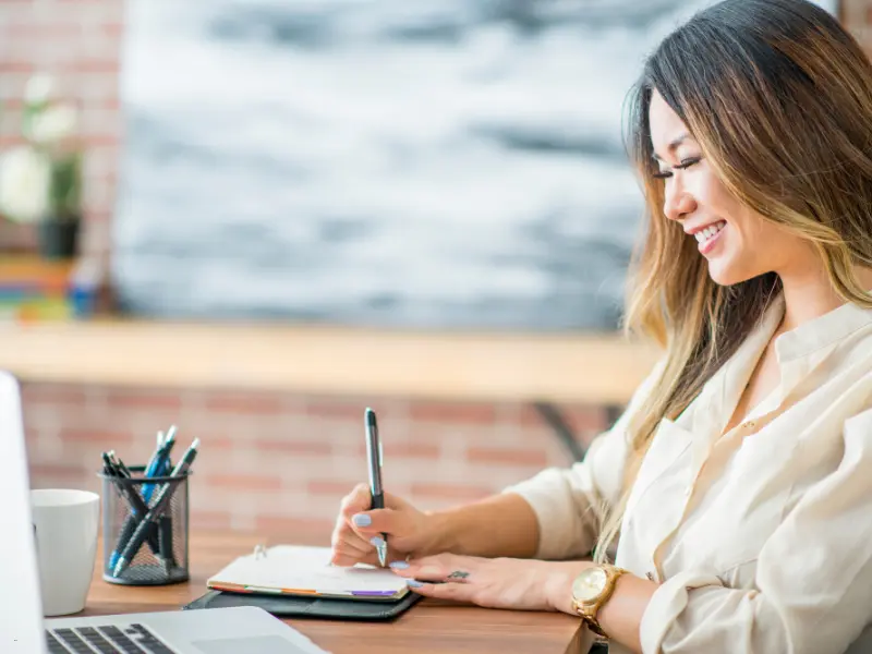 Appointment scheduling software keeps professionals organized, shown by a smiling woman writing in a planner at her desk