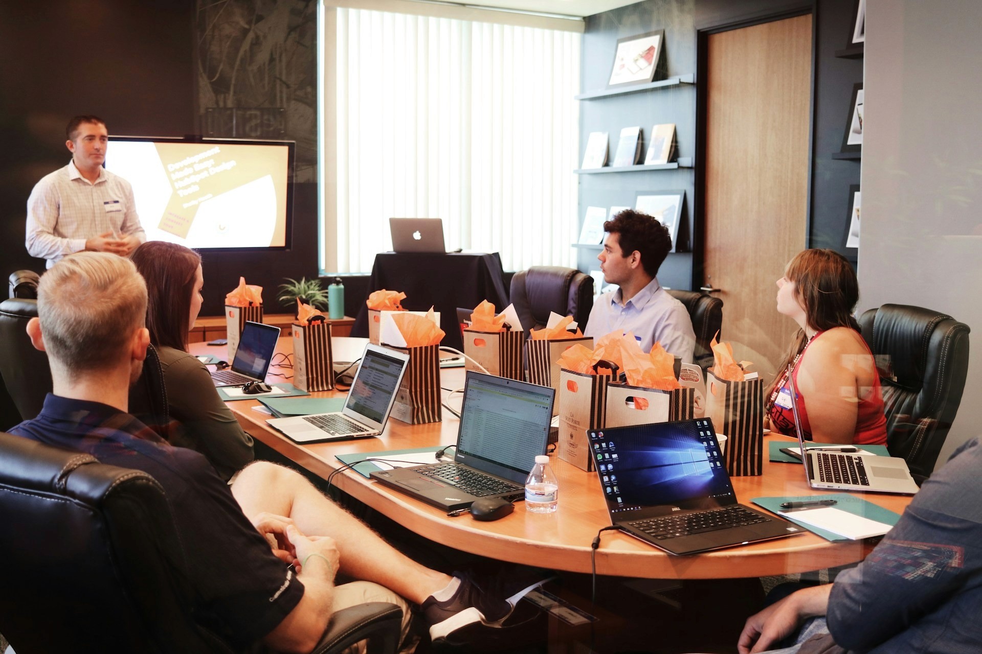 A professional team sitting around a conference table during a presentation, with laptops and gift bags visible on the table.