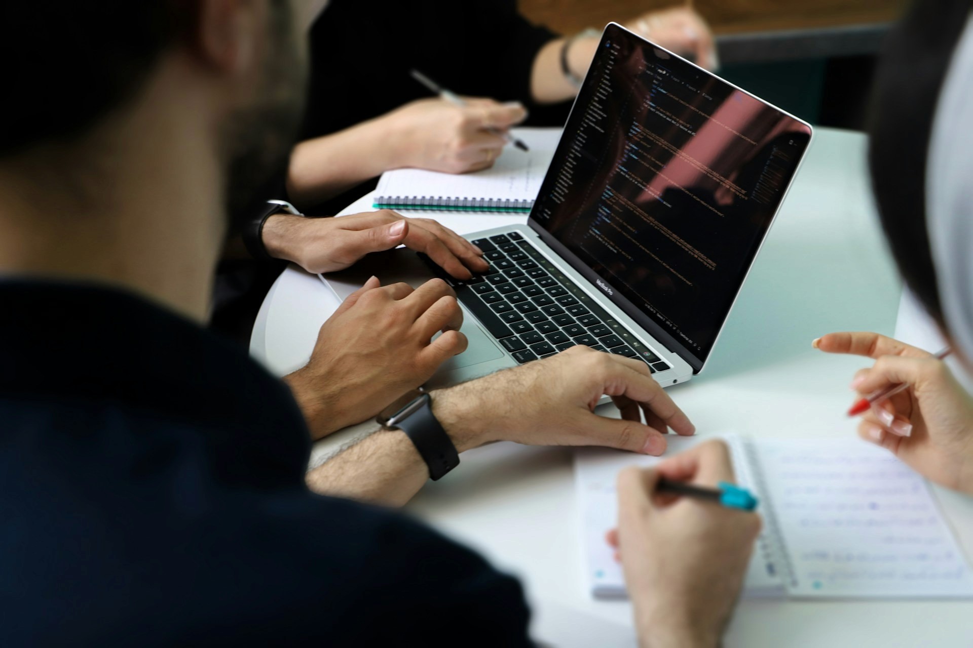 Software developers working together in a technical office environment, reviewing code on a laptop screen.
