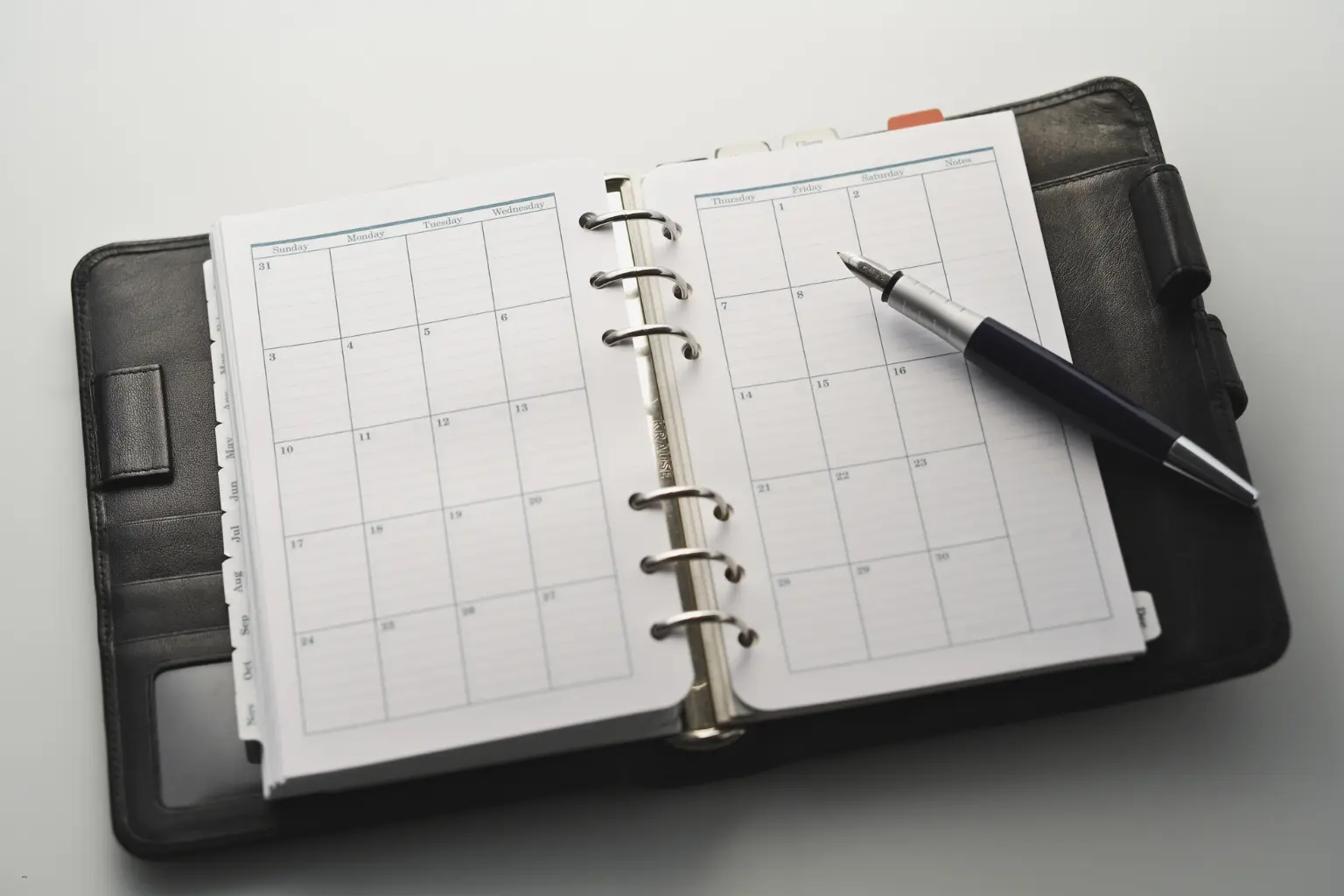 Automated calendar management showing an open leather planner with blank monthly pages and a pen on a white desk