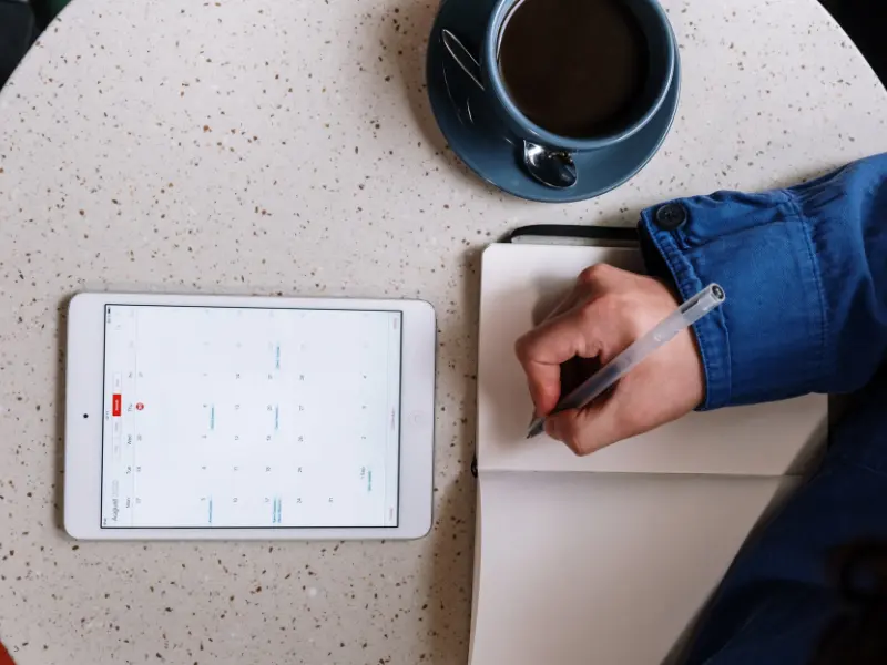 Tools for scheduling include a tablet with a digital calendar and a notebook, as a person takes notes with coffee nearby.