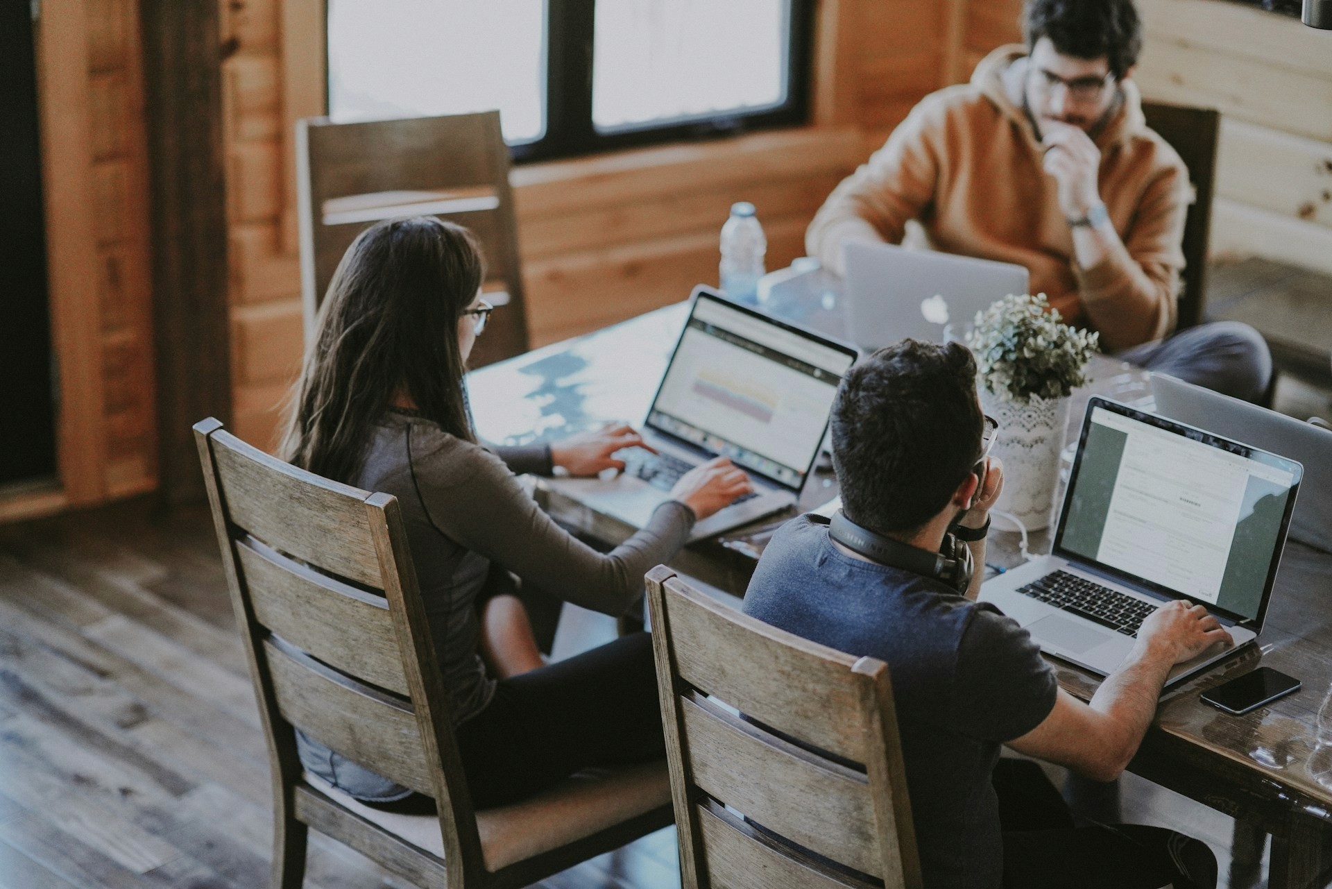 Three professionals working on laptops at a wooden table inside a bright, rustic modern workspace.