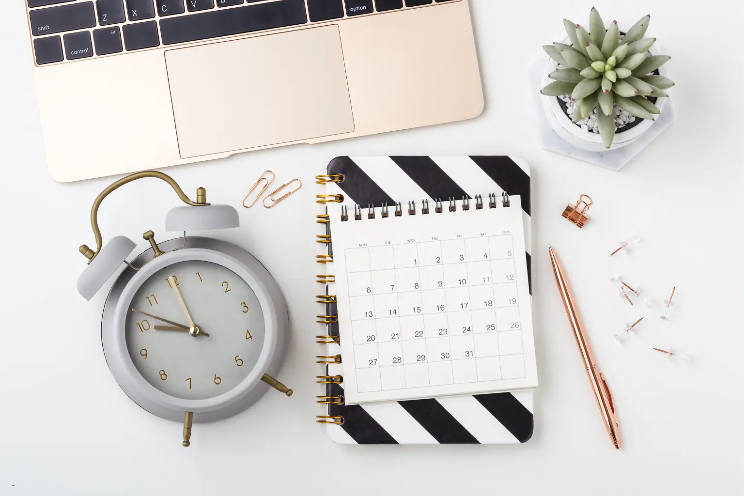 Calendar automation workspace with a laptop, clock, striped notebook calendar, pen, and succulent on a white desk