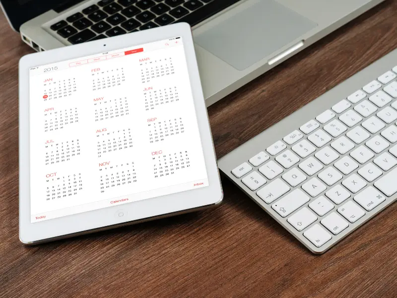 Scheduling software open on a tablet next to a laptop and keyboard on a wooden desk