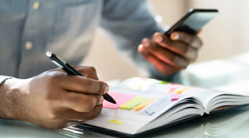 Close-up of hands writing in a planner while holding a phone