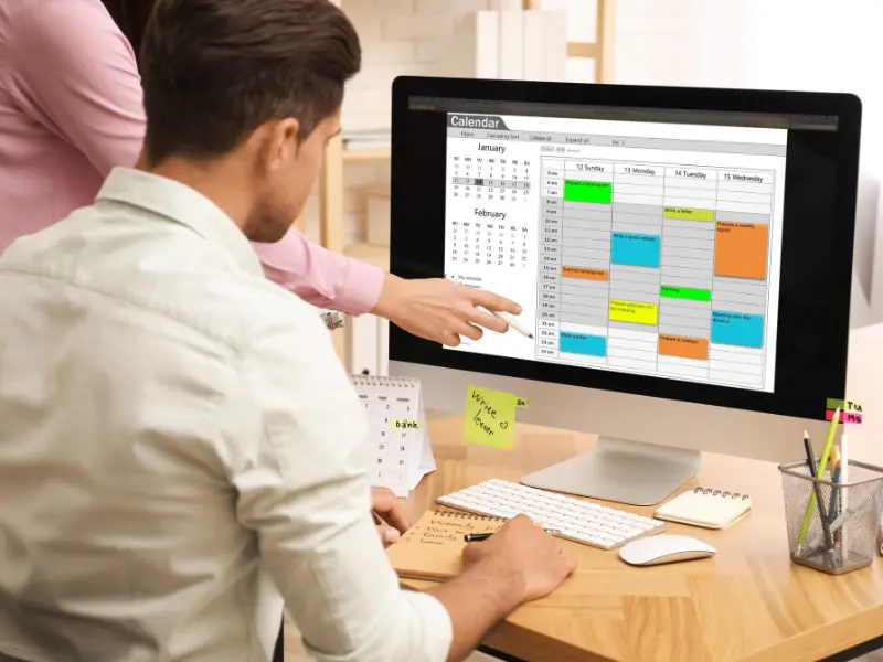 Calendar for team planning shown on a desktop monitor with color-coded events and a colleague pointing at tasks.