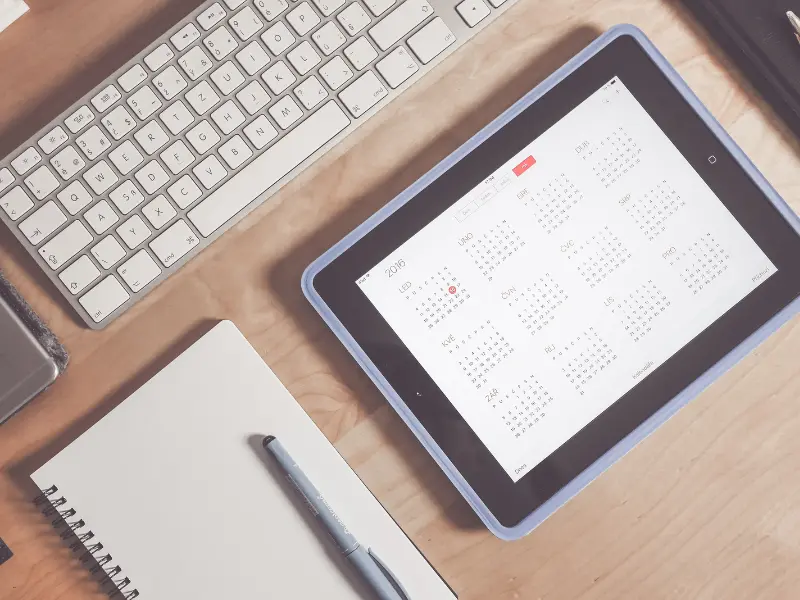 Digital calendar displayed on a tablet, placed on a wooden desk with a keyboard, notebook, and pen