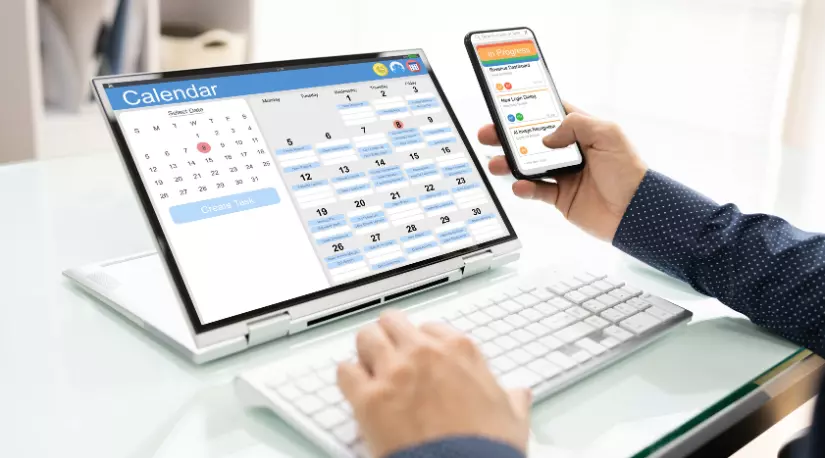Hands using a laptop and phone showing calendar apps