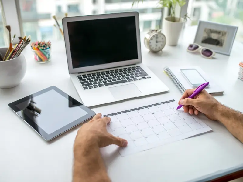 Hands writing on a desk calendar beside a laptop