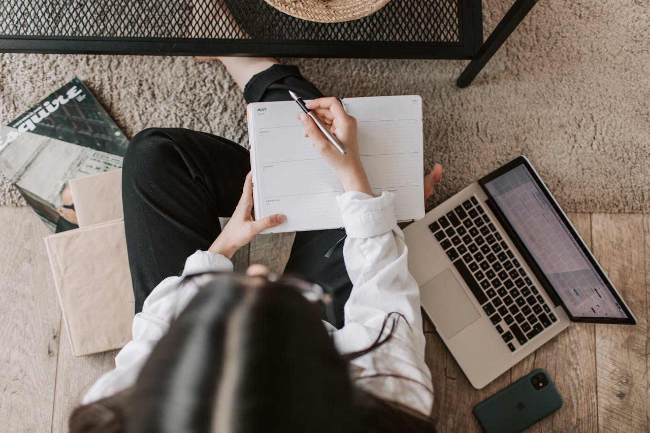 Person writing in a planner beside a laptop